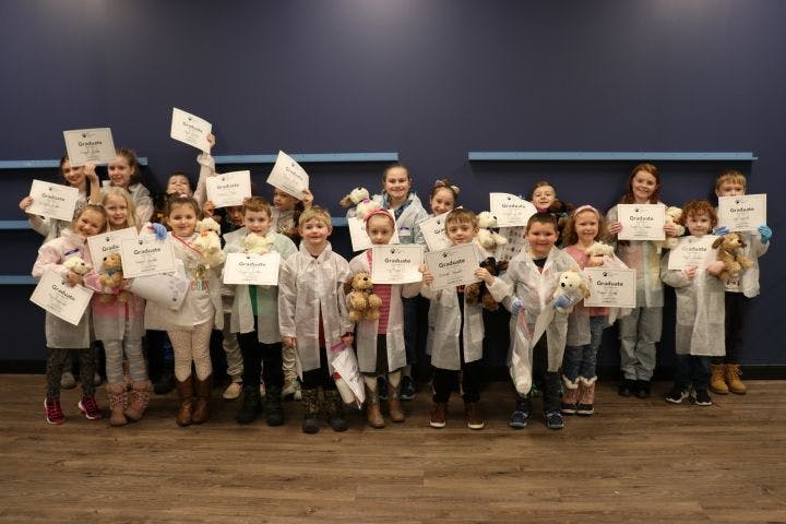 The full class of little veterinarians stands side by side, holding up their diplomas. Each child is in a matching lab coat.