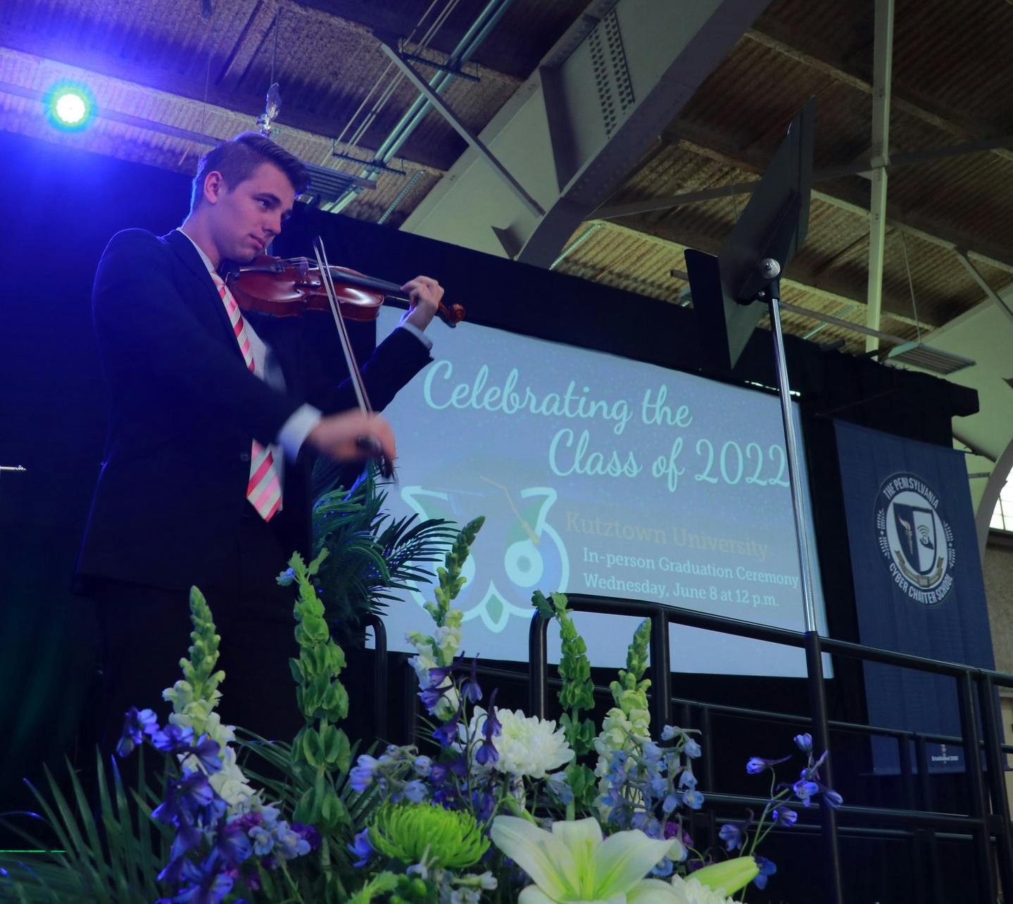 Student in cap and gown plays violin on stage at graduation ceremony.